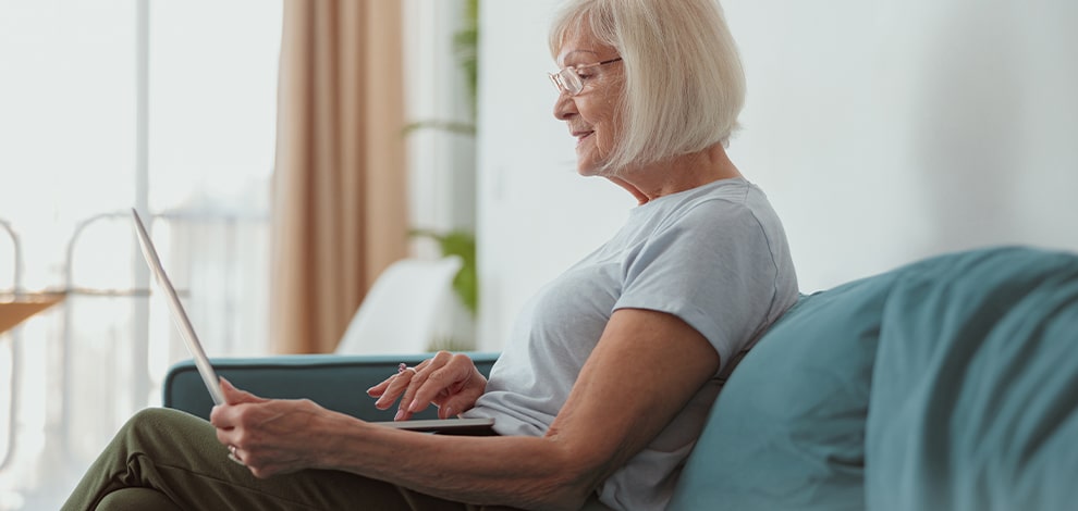Une femme âgée assise confortablement sur un canapé utilise un ordinateur portable. Elle pourrait bénéficier de l'assistance d'Aidant Connect pour naviguer dans ses démarches numériques.