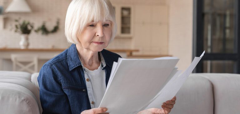 Une femme âgée, assise confortablement dans son salon, lit attentivement des documents. L'expression sérieuse sur son visage suggère qu'elle pourrait être en train de passer en revue ses relevés de pension ou de se renseigner sur le minimum contributif à la retraite.